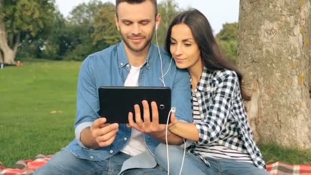 公園の木の近くに座っている若いカップル タブレットと白いヘッドフォンで一緒に音楽を聴いて彼らを抱きかかえた — ストック動画