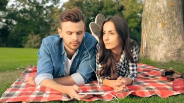 Mutlu genç çift ekose üzerinde yalan, akıllı telefon tutarak ve birlikte müzik kulaklık dinlemek. Park açık havada.