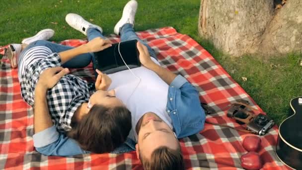 快乐的年轻夫妇躺在格子上 手持数码片 在耳机里听音乐 户外公园 — 图库视频影像