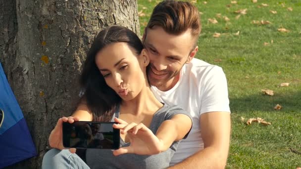 カップルの屋外 木の近くに座っているとスマート フォンの Selfies を撮影します スローモーション — ストック動画