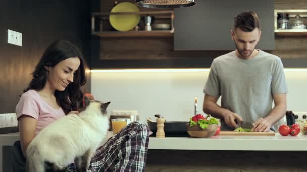 Jovem Feliz Abraçando Gato Segurando Smartphone Enquanto Homem Prepara Salada — Vídeo de Stock