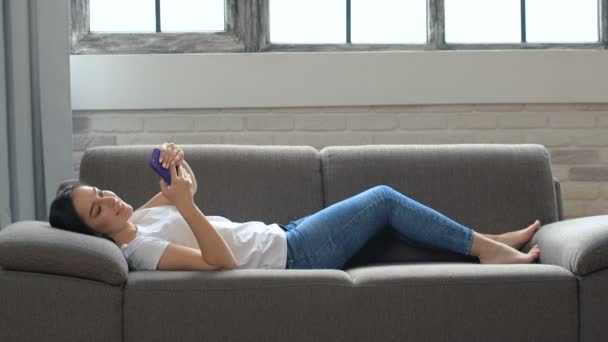 Χαμογελαστός Ευτυχισμένος Νεαρή Γυναίκα Ξαπλωμένη Στον Καναπέ Κρατώντας Smartphone Στα — Αρχείο Βίντεο