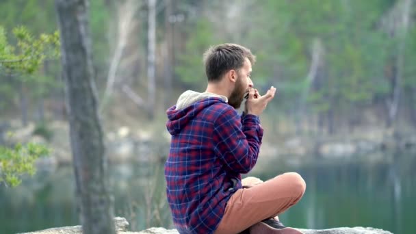 Joven Barbudo Tocando Armónica Sentado Roca Cerca Del Lago Disfrutando — Vídeos de Stock