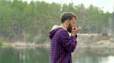 Genç sakal adam armonika açık havada oynuyor. Arka planda orman ile göl.