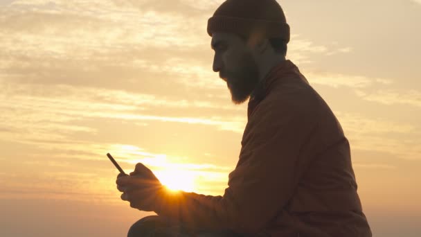 日の出の空の上にスマートフォンを持って座っている男 素敵な太陽の光 スマートフォンを入力して見る — ストック動画