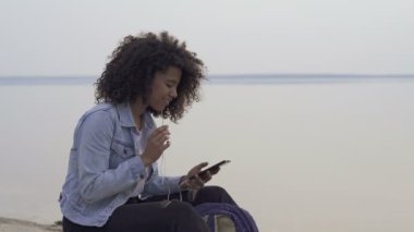 Mutlu afro Amerikalı kadın dışında. Sahilde oturmak, kulaklıkla müzik dinlemek, dinlenmek ve güzel günlerin tadını çıkarmak.