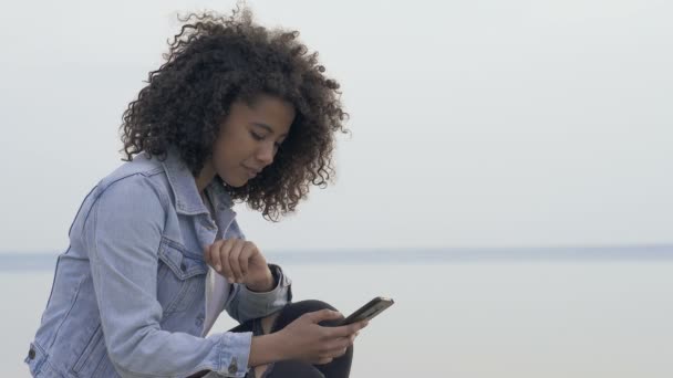 海の近くに座っているアフロアメリカ人女性の肖像画 スマートフォンを持ち タイピングします リラックスして楽しい一日 — ストック動画