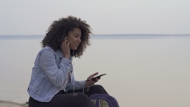 非洲裔美国妇女坐在海边 在耳机里听音乐 享受生活 — 图库视频影像