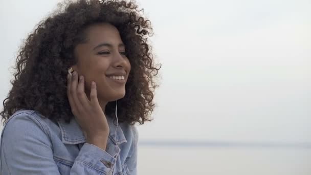 海の近くに座って ヘッドフォンで音楽を聴いて幸せなアフロアメリカ人女性の肖像画をクローズアップ — ストック動画