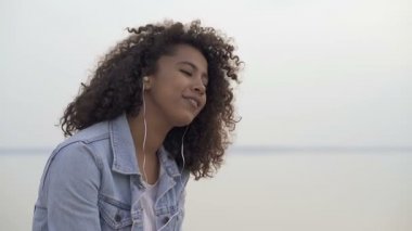 Mutlu afro Amerikalı kadın deniz kenarında oturan. Kulaklıkla müzik dinlemek, dinlenmek ve şarkı söylemek.