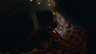 Kıvırcık saçlı afro Amerikan kadın akşam saatlerinde arabanın bagajında oturan. Ekose içinde ısınma. Akıllı telefon tutma ve mesajlaşma.