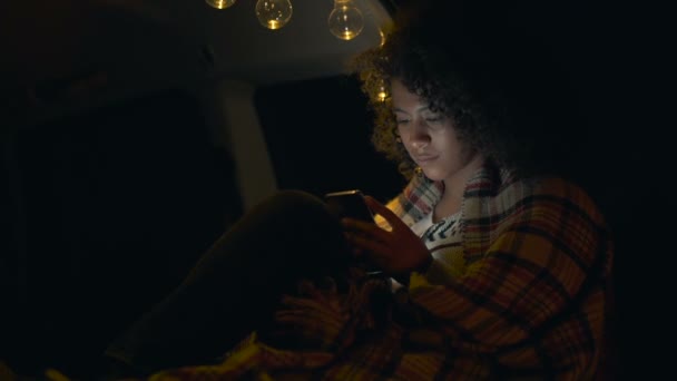 卷曲的头发阿弗罗美国妇女坐在汽车的后备箱在晚上的时间 在格子中热身 手持智能手机和短信 — 图库视频影像