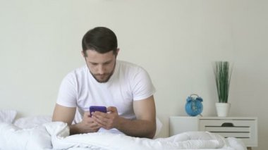 Adam yatakta oturan, akıllı telefon tutan ve ona bakarak.