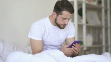 Young man holding smartphone, looking at it and texting. Morning time, sitting at the bed.