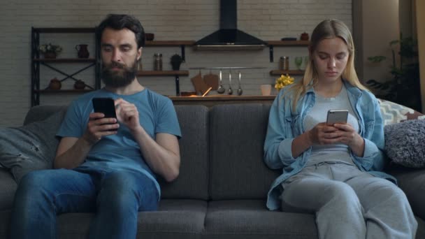 一对年轻夫妇坐在沙发上 不在一起 每个人都拿着智能手机 看着它 沟通和夫妻问题 — 图库视频影像