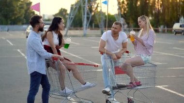 Süpermarket arabaları ile otoparkta duran mutlu arkadaşlar grubu. Erkekler ayakta, kızlar arabada oturuyor. Gülümsüyorlar ve konuşuyorlar..