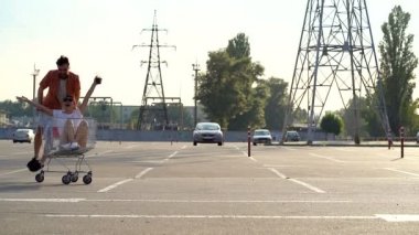 Genç bir çift sakallı adam ve Asyalı kadın park yerinde eğleniyorlar. Süpermarket arabasıyla gezmek. Yavaş çekim.