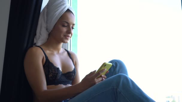 窓に座っている若い女性は 電話でテキストメッセージを書き起こします — ストック動画