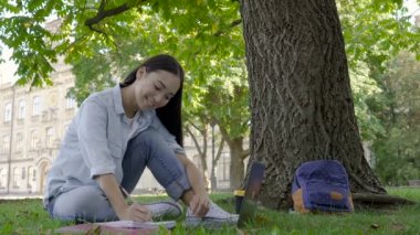 Öğrenci kız dizüstü bilgisayar ile çim üzerinde oturan ve egzersiz kitabında yazma.