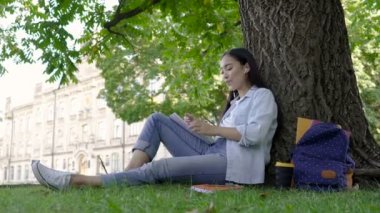 Asiand öğrenci kız parkta ağaç düzgün oturuyor. Kağıt deftere yazma ve kulaklıkla müzik dinleme.
