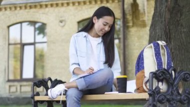 Öğrenci kız üniversite parkı bankta oturan, ders kitabı okuma ve egzersiz kitabında notlar yazma.