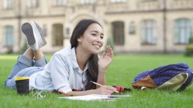 Mutlu ve gülümseyen öğrenci kız parkta çim yatıyor. Kağıt deftere okuyor ve yazıyor..