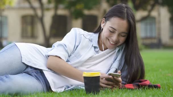 Chica Estudiante Joven Encuentra Césped Parque Sosteniendo Teléfono Inteligente Escuchando — Vídeos de Stock