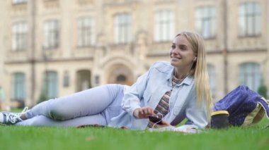 Çimlerin üzerinde yatan mutlu sarışın öğrenci kız. Gülümseyerek, müzik dinleyerek akıllı telefon nad tutarak.