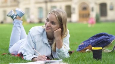 Genç sarışın kadın parkta çimenlerin üzerinde yatıyor ve ders çalışıyor. Rahat. mutlu ve olumlu duygular.