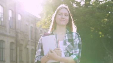 Üniversitenin yanında duran mutlu öğrenci kız. İyi günlerin ve hayatın tadını çıkarıyorum. Ders kitapları tutuyor ve gülümsüyor. El yapımı atış. Slowmotion.