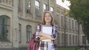 Ders kitaplarını tutan, üniversitenin yanında duran, gülümseyen ve kameraya bakan öğrenci kızın el yapımı fotoğrafı. Slowmotion.