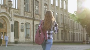 Üniversite binasına yürüyen öğrenci kız. Mutlu ve gülümsüyor, kameraya bakıyor ve ileri doğru yürüyorlar. Arkadan bak. Slowmotion.