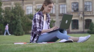 Parkta çimlerin üzerinde oturan genç bir kadın. Dizüstü bilgisayarla çalışmak (serbest çalışma, eğitim). Slowmotion. El çekimi.