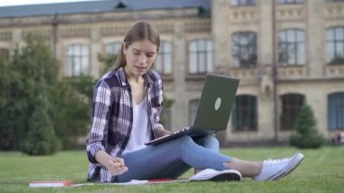 Üniversitenin yakınındaki çimlerin üzerinde oturan kız öğrenci. Dizüstü bilgisayarla çalışmak. Slowmotion.