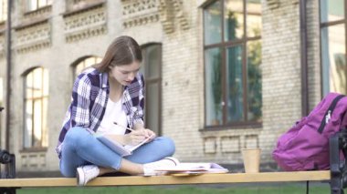 Genç kadın bankta oturuyor, kitap okuyor ve kağıt deftere yazıyor. Eğitim veya serbest çalışma.