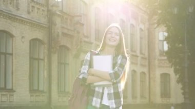 Mutlu ve gülümseyen kadın (öğrenci) üniversite binasıyakınında yürüyüş. Çanta ve tutan ders kitapları ile. Güzel bir yaz günü ve olumlu duygular. El çekimi.