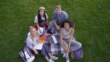 Bir grup öğrenci çimenlerin üzerinde otururken, kameraya bakıyor ve el veda ediyorlar. Üst te. Slowmotion.