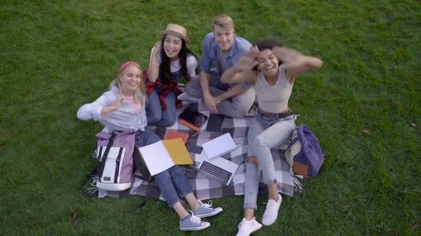 幸せな友人 または学生 のグループは 本やノートを持って草の上に座って カメラを見て 手でやっています トップビュー スローモーション — ストック動画
