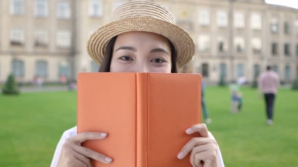 顔の近くに本を持って立っているアジアの若い女性の肖像画をクローズアップ カメラを見てハンドヘルドショット スローモーション — ストック動画