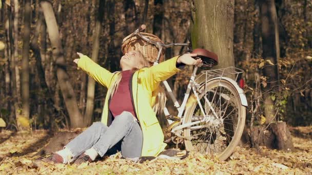 秋天的森林里 迷人的金发女人坐在树旁和自行车旁边 享受生活 树叶落在她身上 慢动作 — 图库视频影像