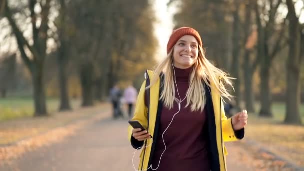 快乐的金发女人在秋天的公园里散步 拿着手机 听着音乐 享受生活 手持射击 慢动作 — 图库视频影像