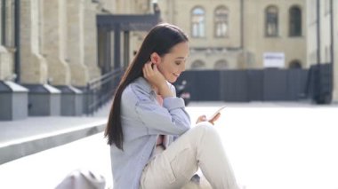 Kaykayın üzerinde oturan, cep telefonu tutan ve müzik dinleyen genç bir kadın. Yavaş çekim.