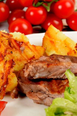 Patateslerin yanında ızgara biftek, yanında salata, domates ve beyaz bir tabakta zeytin.