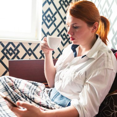 Kızıl saçlı kız kahve fincanı ve cep telefonuyla yatakta oturuyor.