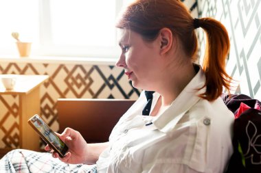 Kızıl saçlı kız yatağında cep telefonuyla oturuyor.