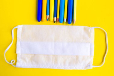 Parlak sarı arka planda renkli kalemler ve tıbbi maske. Okul konseptine geri dönelim. Okul karantina konsepti