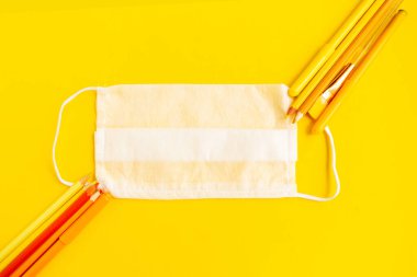 Sarı arka planda renkli kalemler ve tıbbi maske, üst manzara. Okul konseptine geri dönelim. Okul karantina konsepti