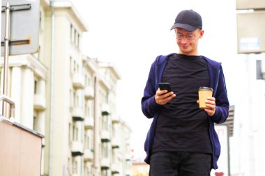Genç bir adam elinde kahve ve telefonla şehir caddesinde yürüyor.
