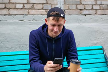 Genç adamın portresi kahve ve telefonla bankta, yakın çekim.