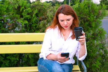 Elinde telefon ve kahve fincanı olan kız parkta bankta oturuyor, kapat.